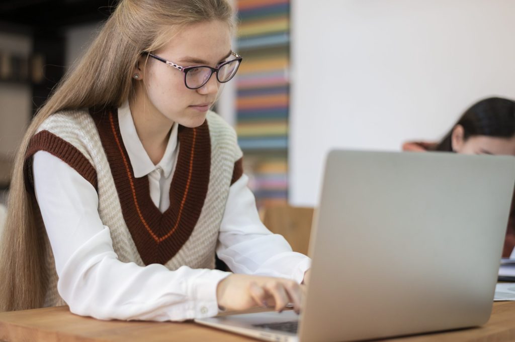 Older female student with glasses sitting at a desk in classroom on a laptop.