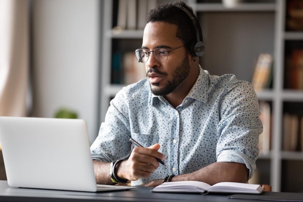 Man with glasses and headphones on sitting at a desk with a laptop in front and a pen and paper.